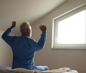 Man sitting on edge of bed stretching and looking out of window